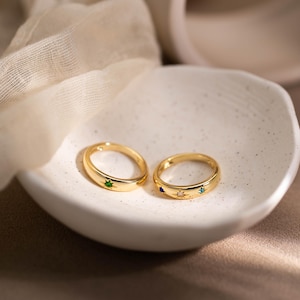 May include: Two gold-toned rings featuring star-shaped settings. One ring displays a single green gemstone, while the other ring features three small embedded gemstones in blue, white, and teal, each arranged within a star-shaped metal engraving.