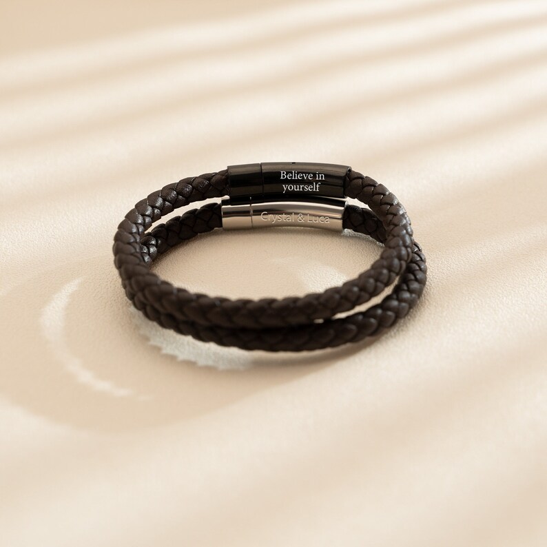 May include: A brown leather bracelet with a braided design and two silver metal clasps. The clasps are engraved with the text "Believe in yourself" and "Crystal & Luca".