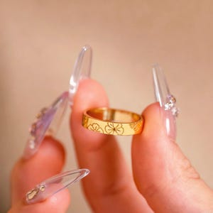May include: A gold-toned metal band ring featuring a delicate, engraved floral pattern that wraps around the exterior surface. The ring has a smooth, polished finish and a consistent width, showcasing detailed line art of small flowers.