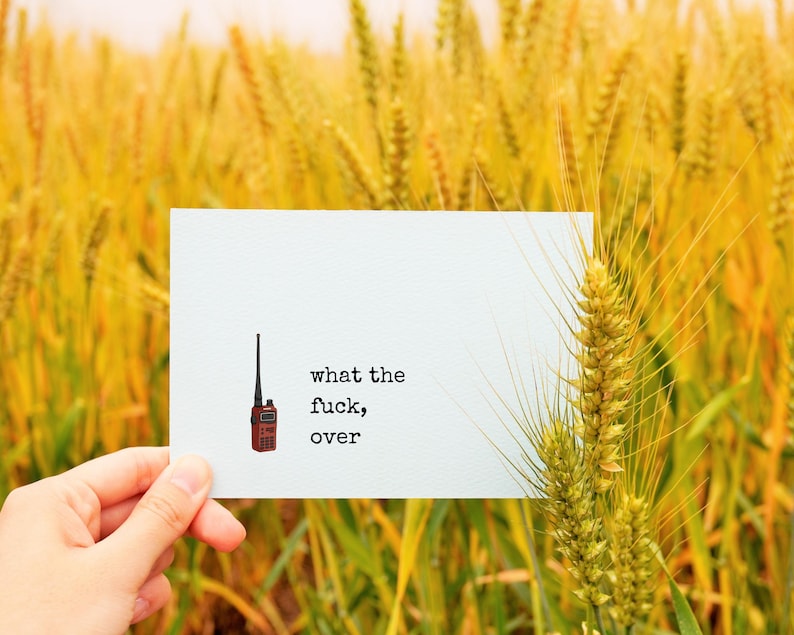 Pu&ograve; includere: Un biglietto bianco con il testo "what the fuck, over" e una piccola illustrazione di un walkie-talkie rosso. Il biglietto &egrave; tenuto davanti a un campo di grano dorato.