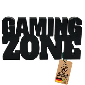 Könnte beinhalten: Ein schwarzes, dreidimensionales Schild mit der Aufschrift "GAMING ZONE" in fetten Blockbuchstaben. Ein kleines, braunes Etikett mit dem Text "DKWRK3D" und einer deutschen Flagge ist angebracht.