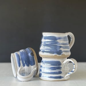 May include: Three ceramic mugs with a white base and horizontal blue brushstroke patterns. One mug is lying on its side, while the other two are stacked vertically. Each mug features a curved handle and a slightly uneven, hand-thrown texture.