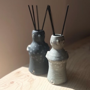 May include: Two ceramic essential oil diffusers in light and dark grey. Each has a rounded shape with a small opening at the top, decorated with white dots and floral accents. Black reeds are inserted into the openings. The diffusers sit on a wooden surface.