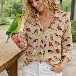 Puede incluir: Camisa beige con botones adornada con un estampado de pájaros vibrante. La camisa presenta diversas ilustraciones de aves en tonos rojos, amarillos, azules y verdes. La modelo lleva pantalones cortos blancos y un collar dorado.