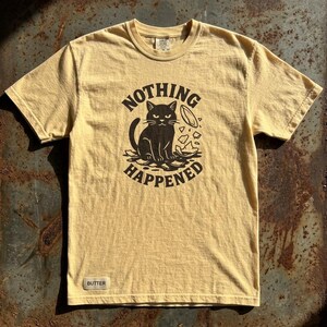 Könnte beinhalten: Eine weiße Keramik-Tasse mit schwarzem Henkel und dem Text "But First, Coffee" in fetter, schwarzer Schrift. Die Tasse steht vor einem verschwommenen Hintergrund, der eine gemütliche Umgebung suggeriert.