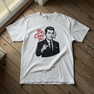Puede incluir: Camiseta blanca con una ilustración en blanco y negro de un hombre con traje fumando un cigarrillo, con el texto rojo "Be Cool Bro". El hombre está señalando. La camiseta está sobre una superficie de madera.
