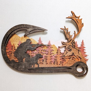 May include: Wooden wall art depicting a hunting scene. A hunter aims a rifle with a dog, and a deer is on the right. The art piece features a layered design with natural wood tones and a mountain and forest backdrop.