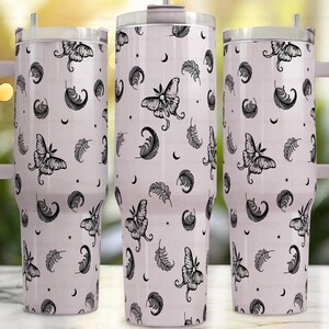May include: A set of three white insulated tumblers with handles, featuring a repeating pattern of black moths, feathers, moons, and stars. The tumblers have a silver lid and a white handle. The design is a whimsical, celestial theme.