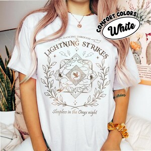 Pu&ograve; includere: T-shirt bianca con un design grafico dorato. Il design include il testo "Dancing Through The Lightning Strikes" e "Sleepless in the Onyx night", fulmini, un drago e un bordo floreale. Sono presenti anche le parole "Comfort Colors" e "White".