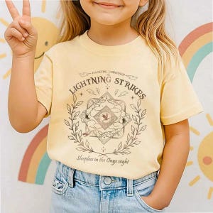 Pu&ograve; includere: Una maglietta giallo chiaro con la scritta "Dancing Through Lightning Strikes" e "Sleepless in the Onyx night". La grafica &egrave; un disegno dettagliato con un emblema centrale, circondato da una corona d'alloro. La maglietta &egrave; abbinata a jeans blu.