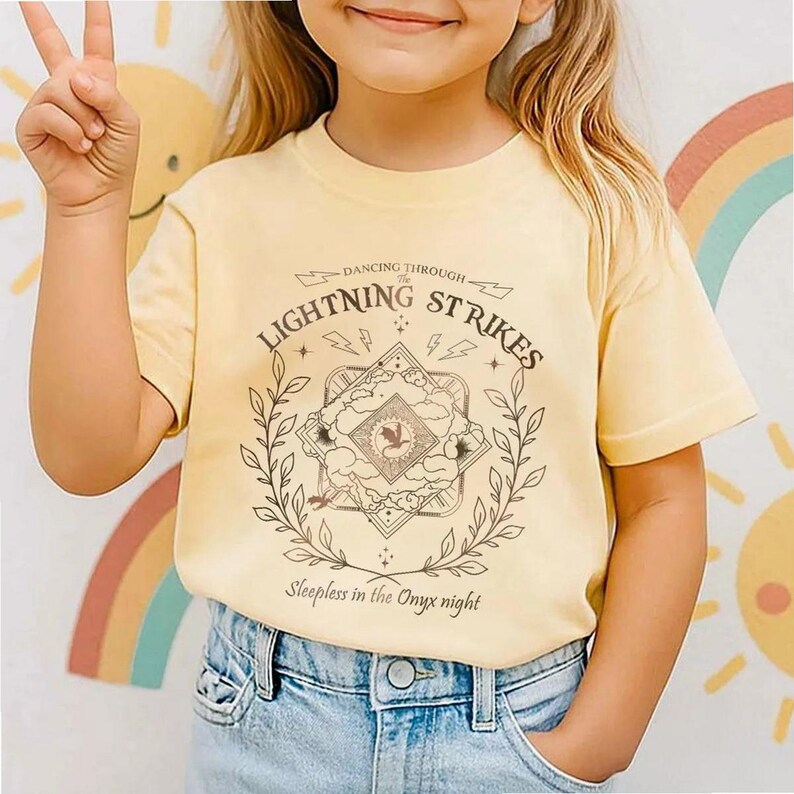 Pu&ograve; includere: Una maglietta giallo chiaro con la scritta "Dancing Through Lightning Strikes" e "Sleepless in the Onyx night". La grafica &egrave; un disegno dettagliato con un emblema centrale, circondato da una corona d'alloro. La maglietta &egrave; abbinata a jeans blu.