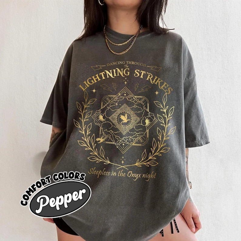 Pu&ograve; includere: T-shirt grigio scuro con un design grafico dorato con il testo "LIGHTNING STRIKES" e "Sleepless in the Onyx night". La maglietta ha anche il testo "COMFORT COLORS Pepper".