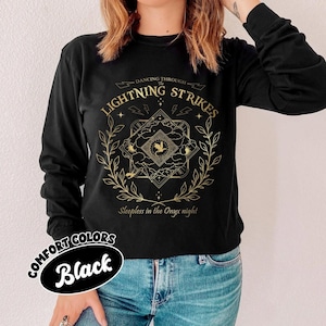 Pu&ograve; includere: Maglietta nera a maniche lunghe con un design grafico dorato. Il design include il testo "Dancing Through The Lightning Strikes" e "Sleepless in the Onyx night", fulmini, un emblema centrale e un bordo floreale. La maglietta &egrave; etichettata "Comfort Colors Black".