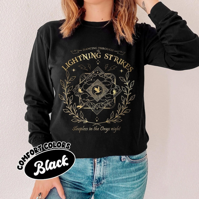 Pu&ograve; includere: Maglietta nera a maniche lunghe con un design grafico dorato. Il design include il testo "Dancing Through The Lightning Strikes" e "Sleepless in the Onyx night", fulmini, un emblema centrale e un bordo floreale. La maglietta &egrave; etichettata "Comfort Colors Black".