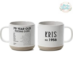 Puede incluir: Taza de cerámica blanca con base beige. Un lado presenta "70 YEAR OLD TEXTING CODE" con abreviaturas y sus significados. El otro lado muestra "KRIS EST. 1956" en negro. Una divertida idea para regalo.