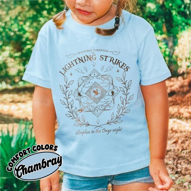 Pu&ograve; includere: T-shirt azzurra con un design grafico. Il design include il testo "LIGHTNING STRIKES" e "Dancing Through" sopra un emblema centrale. L'emblema presenta un uccello e il testo "Sleepless in the Onyx night". La maglietta &egrave; etichettata "Comfort Colors Chambray".