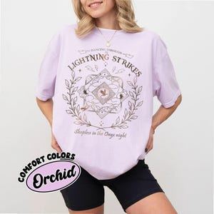 Pu&ograve; includere: Una maglietta lilla con la scritta "Dancing Through Lightning Strikes" e un design grafico. La maglietta presenta anche la scritta "Sleepless in the Onyx night" e la parola "Orchid" in un design circolare.
