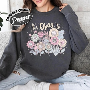 Puede incluir: Sudadera gris oscuro con un diseño floral. El diseño incluye el texto "It's Okay to" sobre un ramo de flores en tonos pastel con frases como "Ask for Help" y "Change Your Mind". La sudadera tiene cuello redondo y mangas largas.
