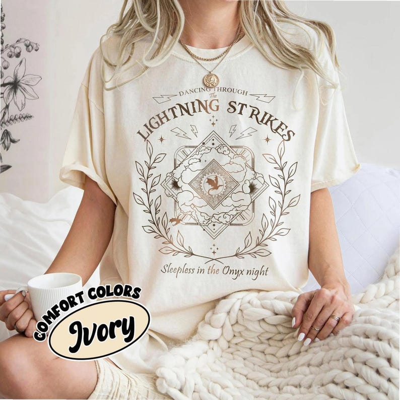 Pu&ograve; includere: T-shirt color avorio con un design grafico. Il design include il testo "Dancing Through The Lightning Strikes" e "Sleepless in the Onyx night." La grafica presenta un emblema centrale con un uccello ed elementi floreali.