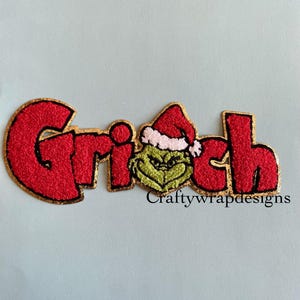 May include: Embroidered patch featuring the word "Grinch" in red, outlined in gold. The "i" is replaced with a green Grinch face wearing a Santa hat. The text "Craftywrapdesigns" is below the word.