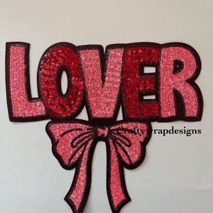 May include: A pink and red sequined iron-on patch with the word "LOVER" in large letters and a pink bow underneath.