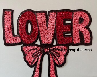 Peut inclure: Un patch thermocollant en sequins rose et rouge avec le mot "LOVER" en grandes lettres et un nœud rose en dessous.