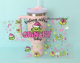 Pode incluir: Um copo térmico rosa com tampa e alça brancas, com um gráfico do Grinch usando um gorro de Papai Noel e as palavras "Feeling Extra Grinchy Today". Adesivos decorativos com rostos do Grinch, sinos e bengalas doces circundam o copo.