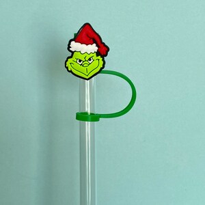 K&ouml;nnte beinhalten: Ein gr&uuml;ner Grinch-Charakter mit einem roten Weihnachtsmann-Hut ist an einem klaren Glasstrohhalm mit einem gr&uuml;nen Silikon-Strohhalmhalter befestigt.