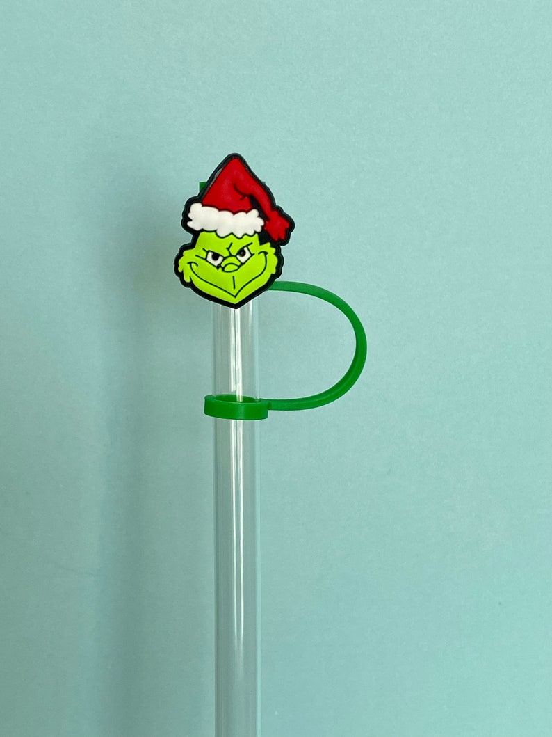 K&ouml;nnte beinhalten: Ein gr&uuml;ner Grinch-Charakter mit einem roten Weihnachtsmann-Hut ist an einem klaren Glasstrohhalm mit einem gr&uuml;nen Silikon-Strohhalmhalter befestigt.