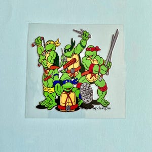 May include: A sticker depicting the Teenage Mutant Ninja Turtles in dynamic poses. Each turtle is a different colour, with weapons and gear. The sticker has the text "fanywrapdesigns" at the bottom.