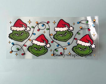 Pode incluir: Uma folha de autocolantes transparente com três personagens de desenhos animados verdes com gorros de Pai Natal vermelhos e brancos, ligados por um fio de luzes de Natal coloridas e pequenas estrelas amarelas, com o texto Craftywrapdesigns impresso.