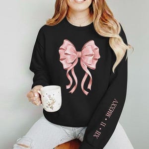 Puede incluir: Sudadera negra con un gran lazo rosa en la parte delantera. El lazo tiene sombreado detallado y cintas fluidas. La manga tiene los números romanos XII • II • MMXXV impresos en rosa. Se sostiene una taza blanca con diseño floral.