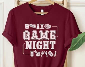 May include: A maroon short-sleeved t-shirt featuring white graphic text that reads GAME NIGHT in the center. The design is surrounded by small white icons of playing cards, dice, a pawn, an hourglass, a dartboard, wine glasses, and chips.