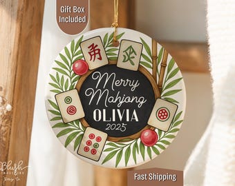 May include: A round, white ceramic ornament with a black center featuring the text "Merry Mahjong OLIVIA 2025." The ornament is decorated with Mahjong tiles, red spheres, and green foliage. A gold string is attached for hanging.