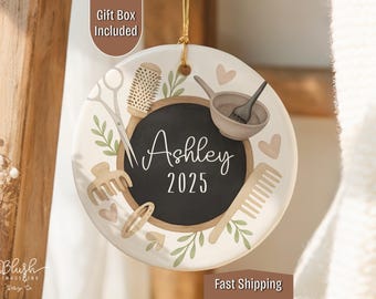 May include: A round, white ceramic ornament with a gold string for hanging. It features a black chalkboard center with the name "Ashley" and the year "2025" in white script. The design includes hair styling tools and decorative elements.