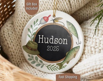 May include: A round, white ceramic ornament with a fishing theme. It features a black circle with the name "Hudson" and the year "2025". The ornament is decorated with fishing lures, a hat, and fish illustrations. A gold string is attached for hanging.