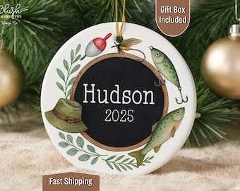 May include: A white ceramic ornament with a black center circle featuring the name "Hudson" and the year "2025". The ornament is decorated with fishing-themed illustrations, including a hat, fish, and fishing lures. A gold string is attached for hanging.