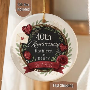 May include: A round, white ceramic ornament with a black center, celebrating a 40th anniversary. It features a burgundy ribbon, gemstones, a rose, and the names Kathleen & Henry, with the date 02.14.2026. A gold string is attached for hanging.