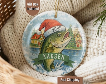 May include: A round Christmas ornament featuring a cartoon bass wearing a Santa hat with the year 2025. The ornament has a winter scene with a lake, houses, and a Christmas tree. The name "Karsen" is on a banner.
