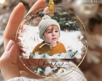 May include: A round glass Christmas ornament with a photo of a child wearing a white hat and a yellow sweater. The ornament has a gold trim and the text "2024 Charlie" is printed on the bottom.