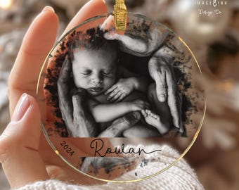 Puede incluir: Una bola de Navidad redonda y transparente con una foto en blanco y negro de un recién nacido en manos. El adorno tiene un borde dorado y una cinta dorada para colgar. El nombre "Rowan" y el año "2024" están impresos.