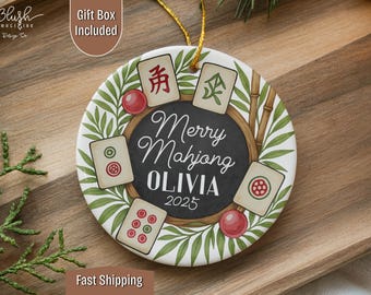 May include: Round ceramic ornament with a Mahjong theme. The ornament features a black center with the words "Merry Mahjong OLIVIA 2025". It is decorated with Mahjong tiles, red spheres, and green bamboo leaves. A gold string is attached for hanging.