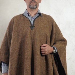 Pode incluir: Um poncho marrom com bainha com franjas. O poncho tem decote em V com um detalhe em couro. A pessoa na imagem está usando uma boina preta e uma camisa com gola por baixo do poncho.