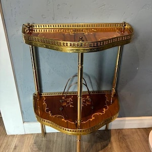 May include: A vintage, two-tiered side table with a half-moon shape. The table features a dark wood surface with inlaid floral designs, accented by a gold-toned metal frame and legs. The table measures approximately 30 inches high.