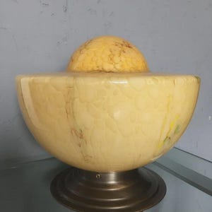 Può includere: Una lampada vintage in stile Art Déco con un paralume a forma di ciotola color crema e una parte superiore più piccola e arrotondata. Il paralume presenta un motivo screziato e poggia su una base color bronzo. Apparecchio di illuminazione decorativo.