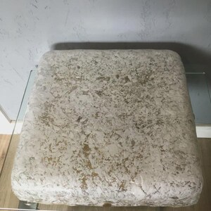 Könnte beinhalten: Ein quadratischer, gepolsterter Hocker mit einer strukturierten, cremefarbenen und goldenen Oberfläche. Der Hocker steht auf einem Tisch aus klarem Glas mit einem silbernen Metallrahmen. Die Oberfläche des Hockers hat ein gesprenkeltes Aussehen. Ca. 60 cm x 60 cm.