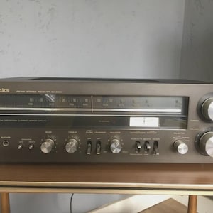 以下が含まれることがあります： ダークグレー仕上げのヴィンテージTechnics SA-300レシーバー。チューナーダイヤル、さまざまなコントロールノブ、電源ボタンを備えています。フロントパネルにはTechnicsのロゴとモデル情報が表示されています。