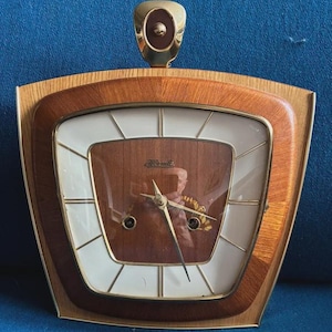 Könnte beinhalten: Eine Vintage-Holzuhr mit hellbraunem Rahmen und dunkelbraunem Zifferblatt. Die Uhr hat einen goldfarbenen Griff oben und goldfarbene Zeiger. Das Zifferblatt hat ein weißes und braunes Design mit goldfarbenen Akzenten.