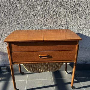 Op de afbeelding: Een houten naaistafel met een rechthoekig blad en een gedeeld deksel. Het meubelstuk is voorzien van een horizontale lade met een geïntegreerde ovale handgreep en een daaronder hangende mand van gevlochten riet op vier zwenkwielen.