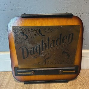 May include: A vintage wooden sign with a brass plate displaying the word "Dagbladen". The sign has a dark brown frame and a lighter wood backing. The brass plate features etched floral designs. The sign is designed to be hung.